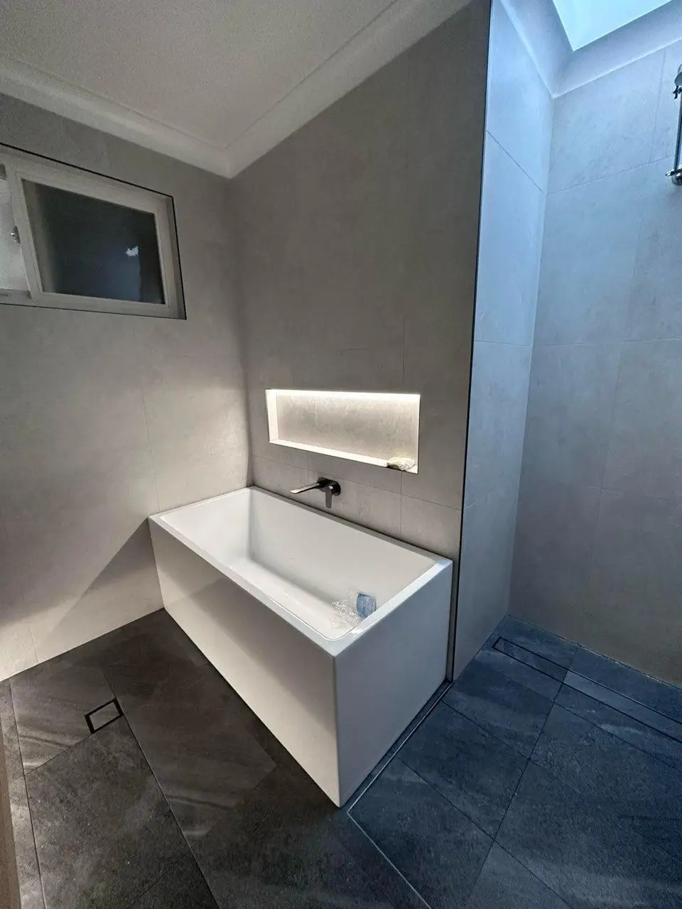 Renovated bathroom with a back to wall bath, backlit tiled niche, large format grey wall tiles and a linear floor waste
