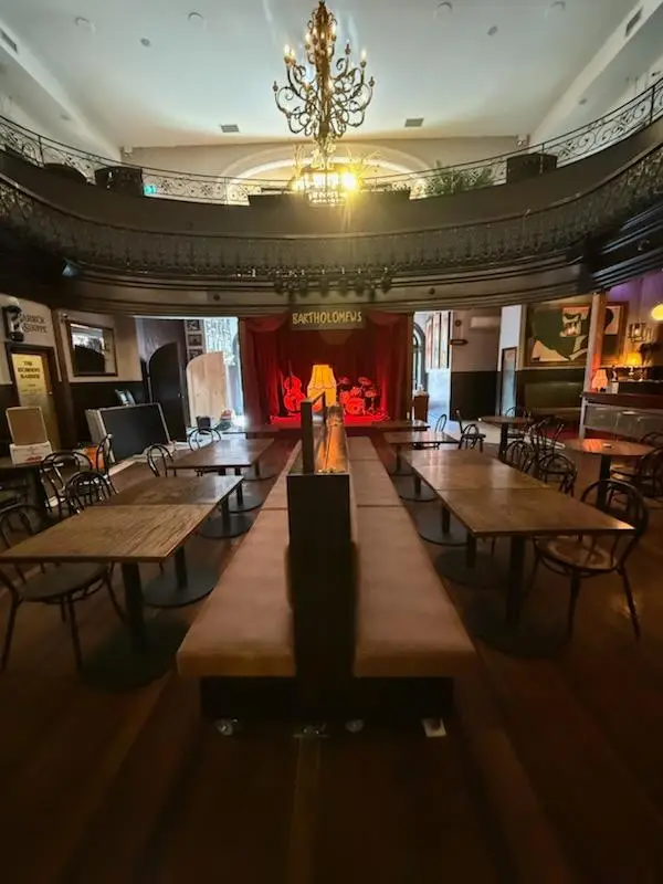 Pub function room with long communal tables, a central banquette spine and a stage under a chandelier