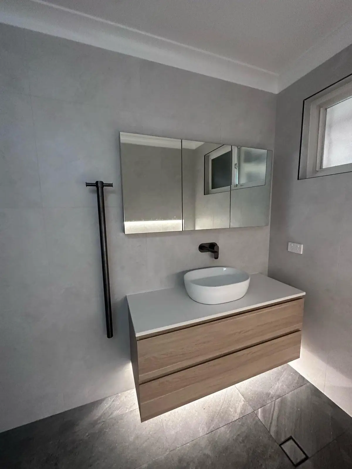 Wall hung vanity in a timber finish with a vessel basin, wall mounted black mixer and a three door mirrored shaving cabinet