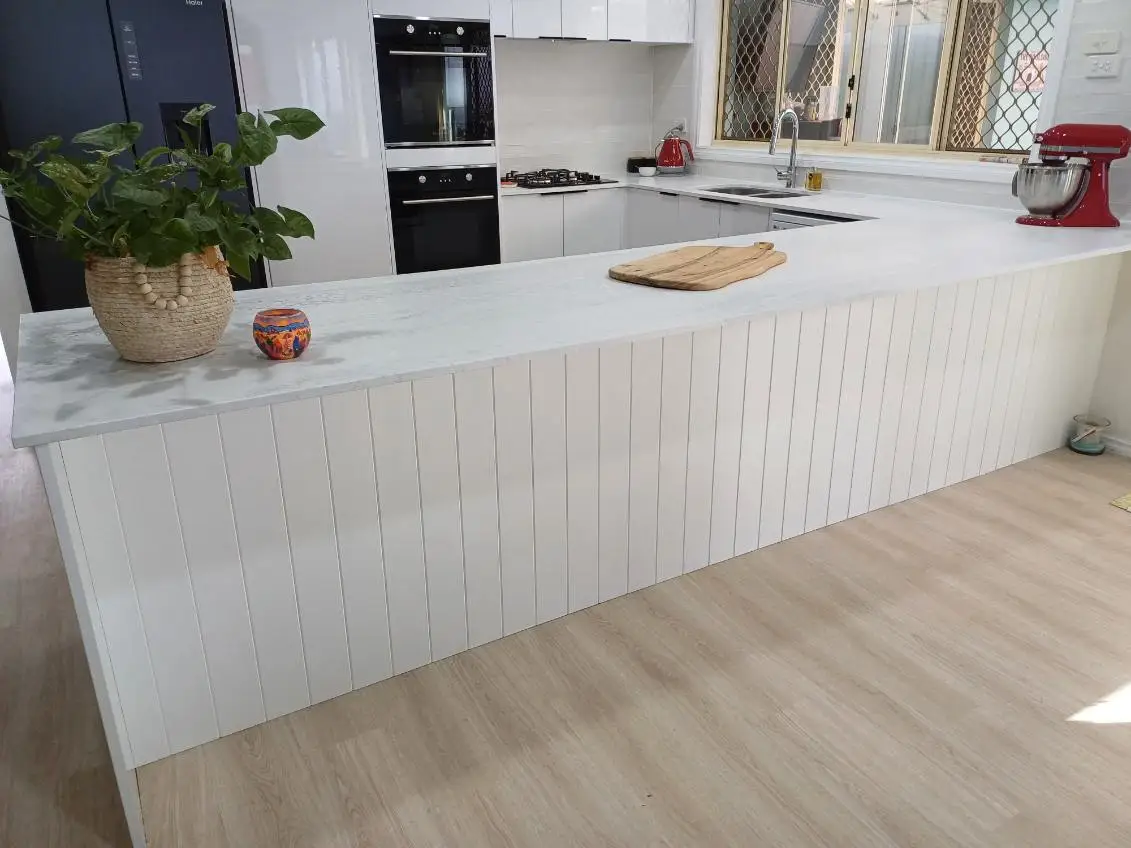 Kitchen peninsula finished in white VJ panelling under a marble-look benchtop, looking back to the cooktop and oven run